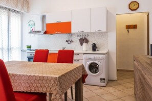 A compact kitchen with a washing machine, a dining table with chairs, and a clock on the wall.