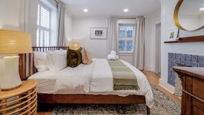 Hotelkamer met een bed, een bedlampje, een raam met gordijnen, een lijst met een afbeelding aan de muur, en een ronde spiegel boven een houten dressoir.