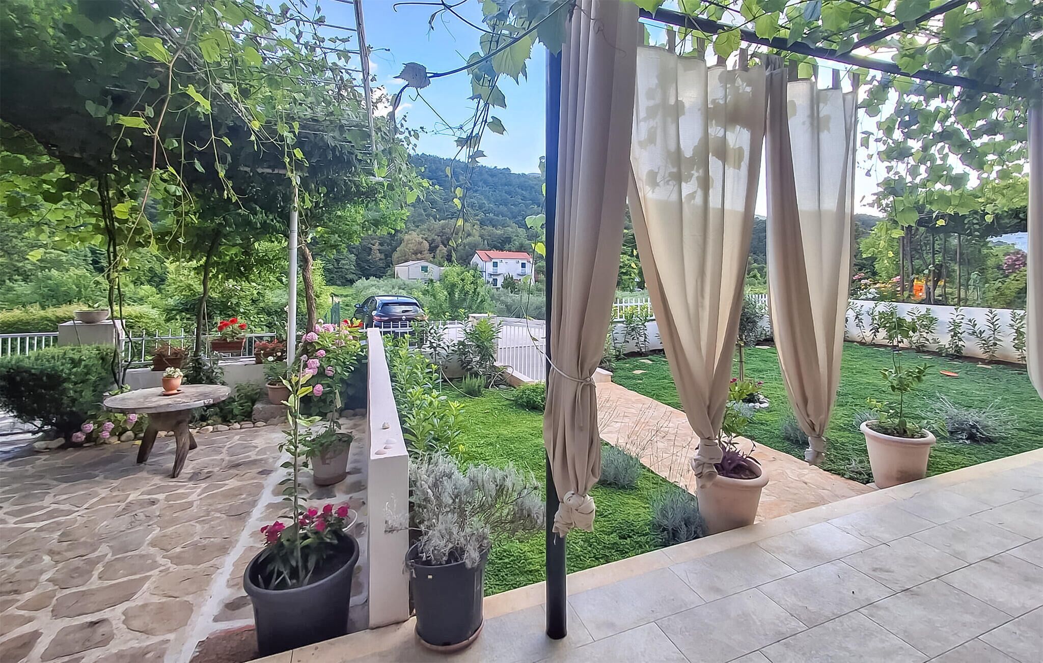 A patio with a stone floor, potted plants, a table, and a view of a green garden and distant buildings.