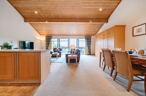 A spacious living area with a wooden ceiling, a dining table set for four, and a kitchen area with wooden cabinets and a flat-screen TV.