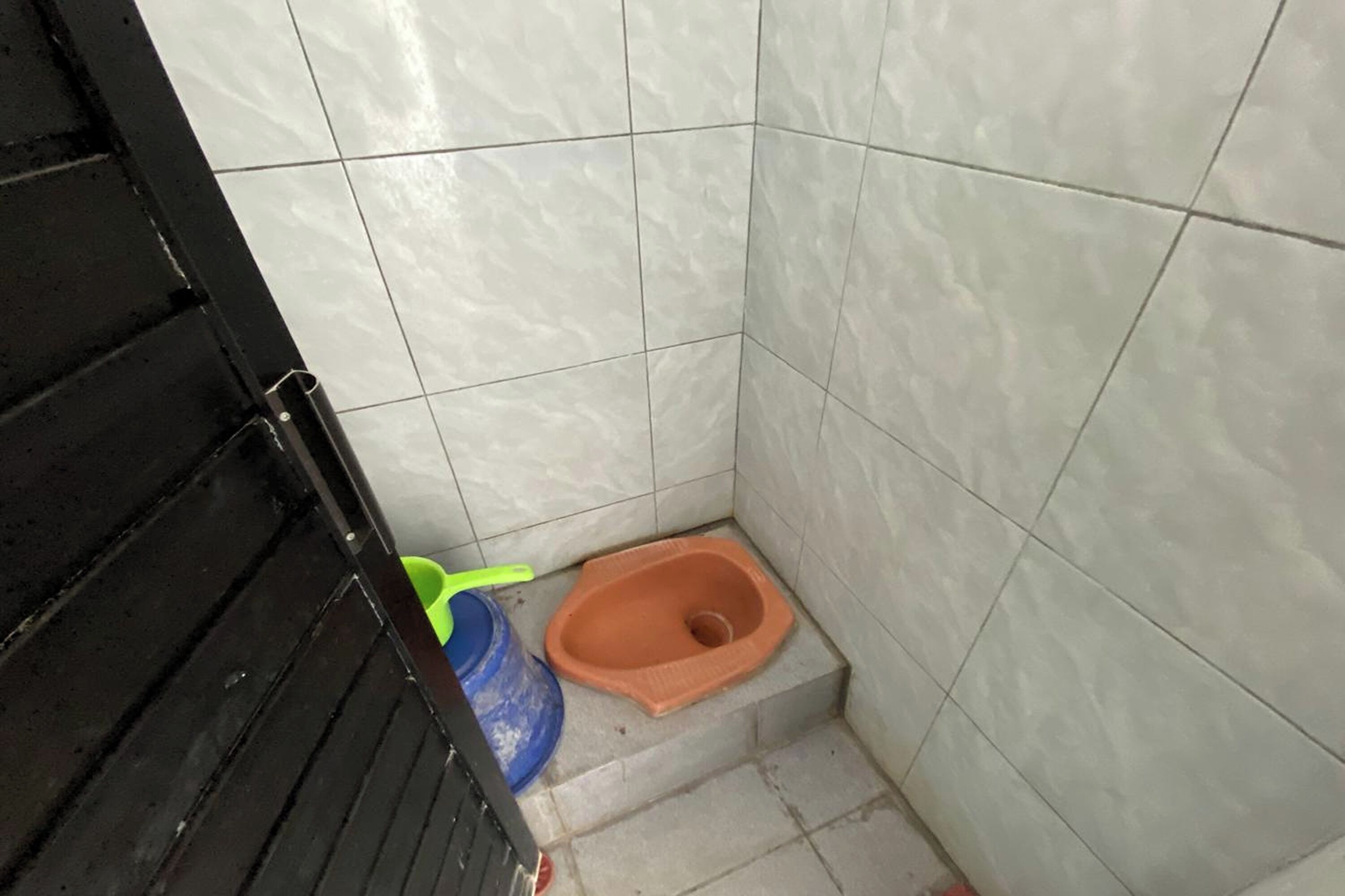 Pieni, laatoitettu kylpyhuone, jossa on wc-istuin, sininen ja vihreä ämpäri sekä musta ovi.