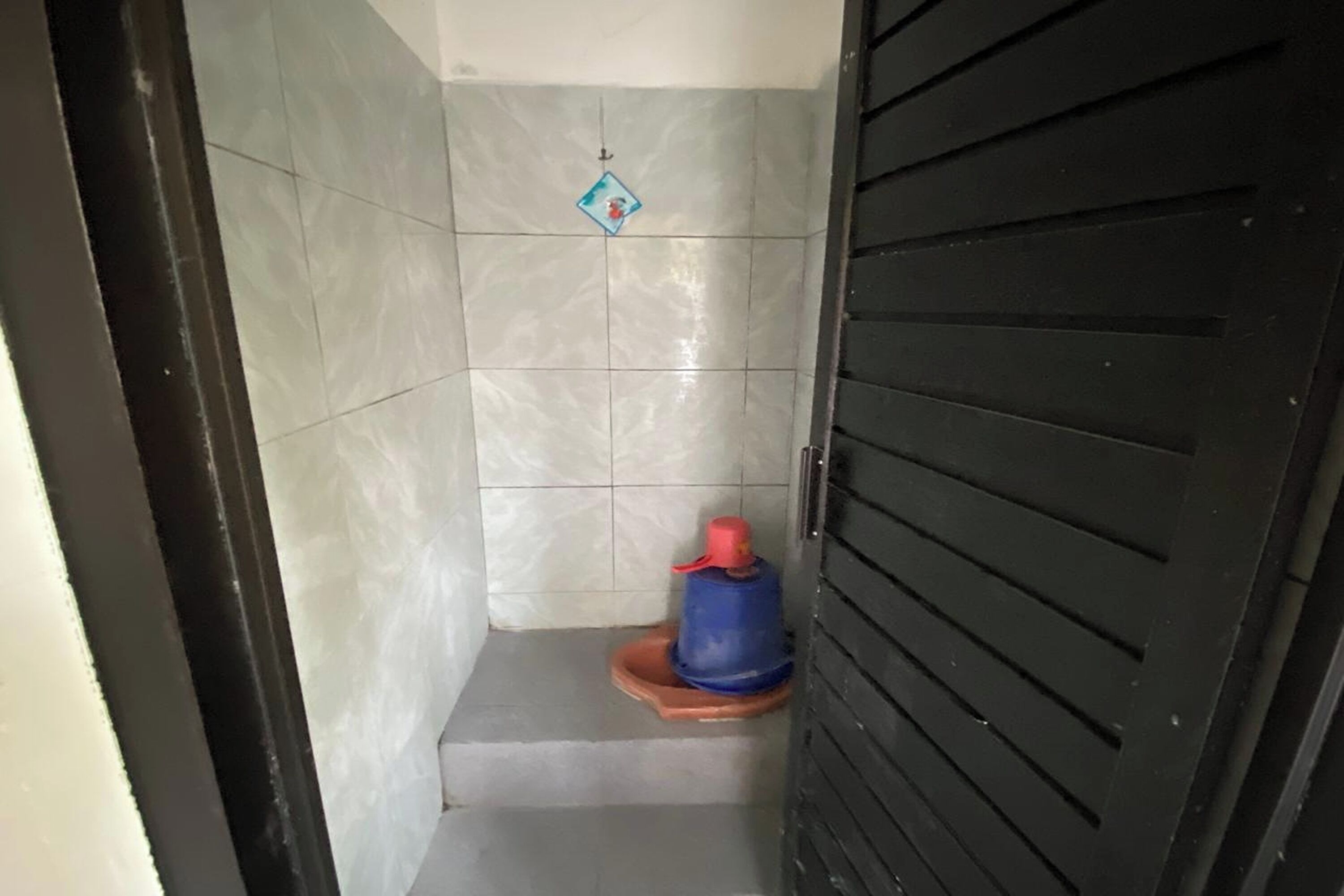 Een kleine, afgesloten ruimte met betegelde wanden, een blauw en rood object op de vloer, en een zwarte deur.