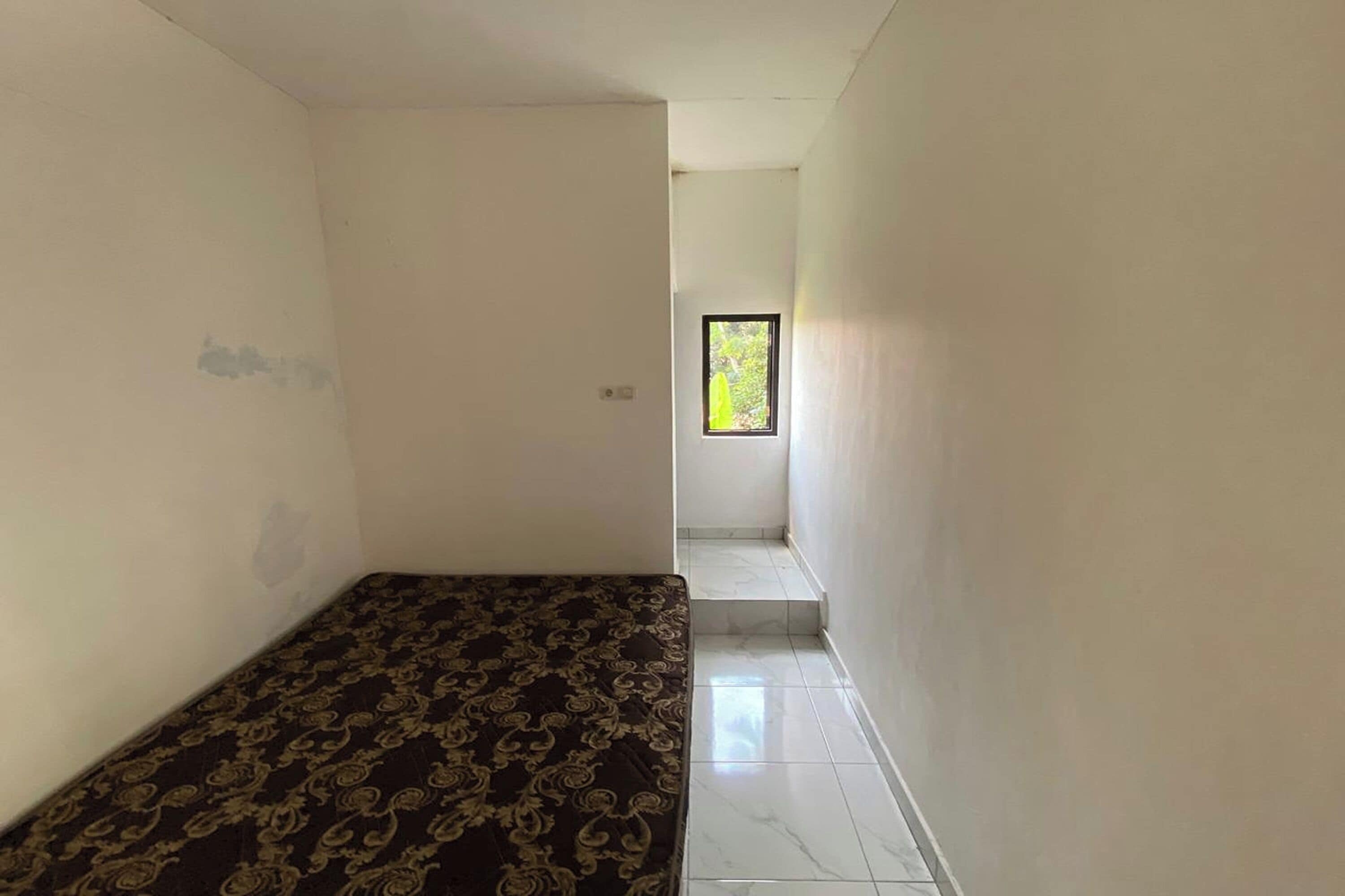 A narrow hallway with a patterned bed at the end, leading to a small window with a view of greenery.