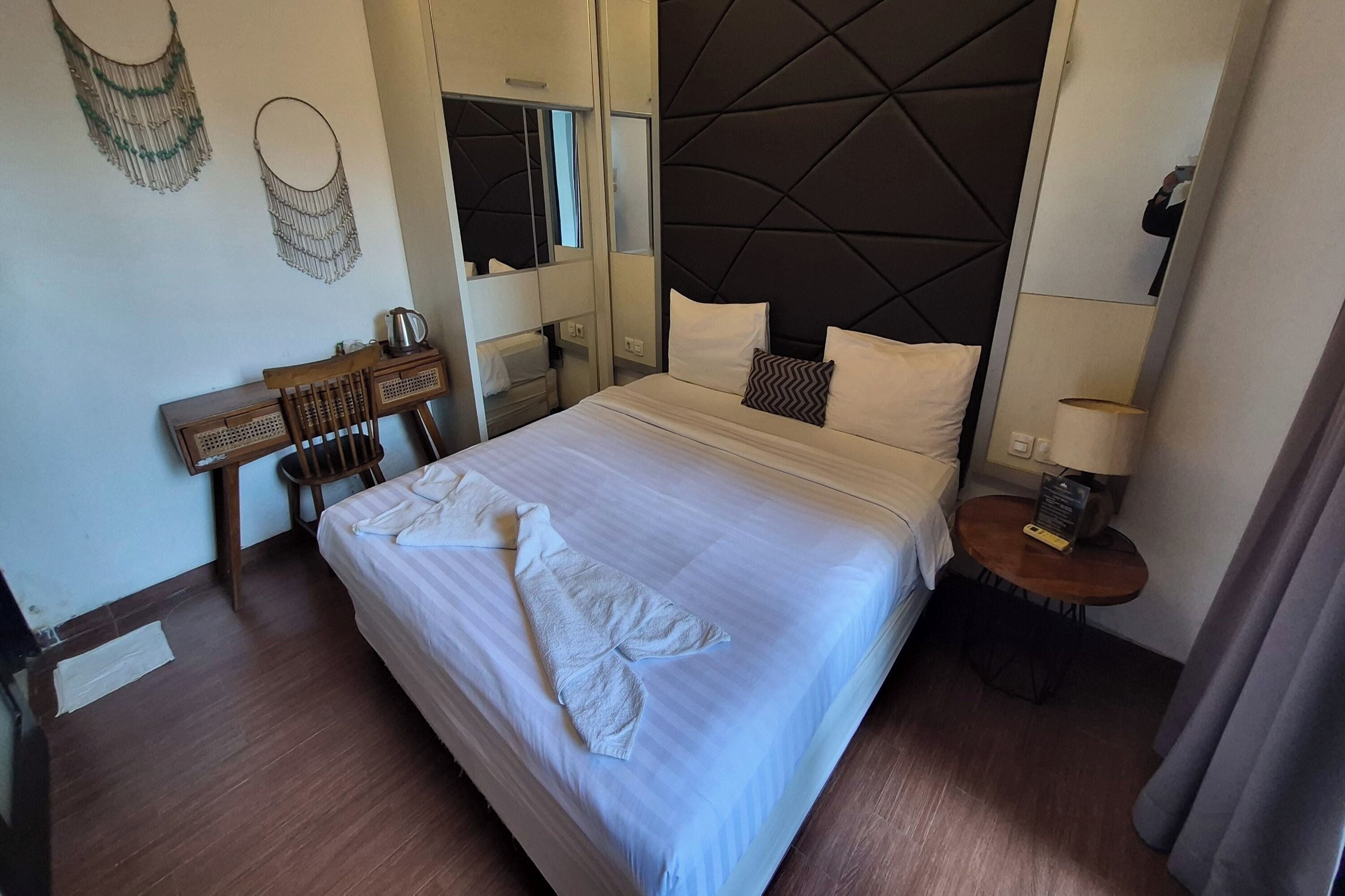 A hotel room with a bed, a desk, a chair, a bedside table, and a mirror.