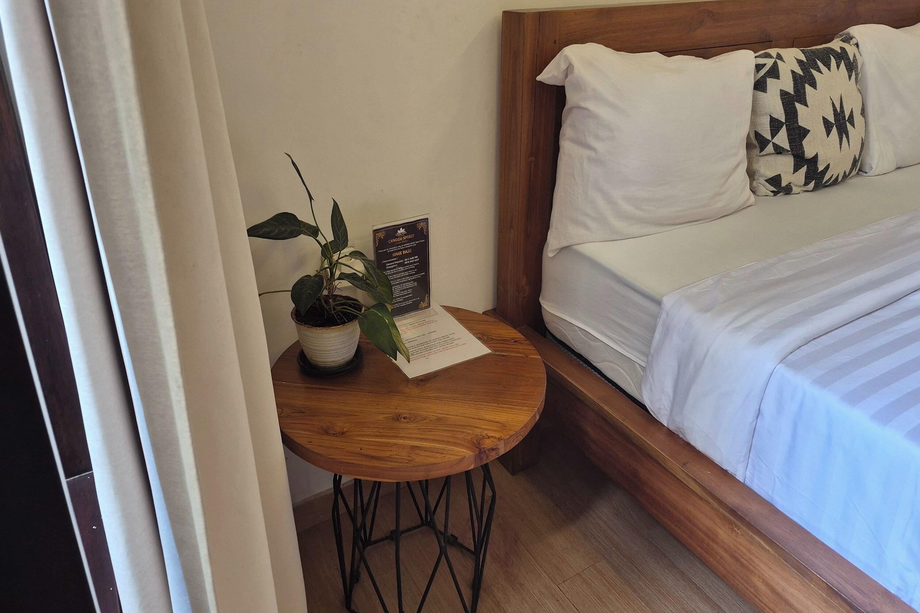 A neatly made bed with white bedding and a black and white patterned pillow. A small wooden table with a potted plant and a house rules sign is positioned beside the bed.