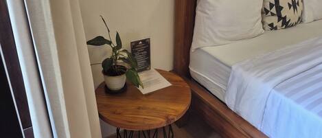 A neatly made bed with white bedding and a black and white patterned pillow. A small wooden table with a potted plant and a house rules sign is positioned beside the bed.