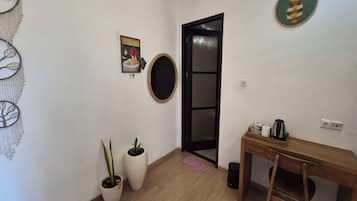 Una habitación con piso de madera, un escritorio con silla, un espejo, una puerta con panel de vidrio y dos plantas en macetas.
