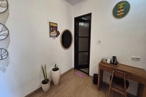 Una habitación con piso de madera, un escritorio con silla, un espejo, una puerta con panel de vidrio y dos plantas en macetas.