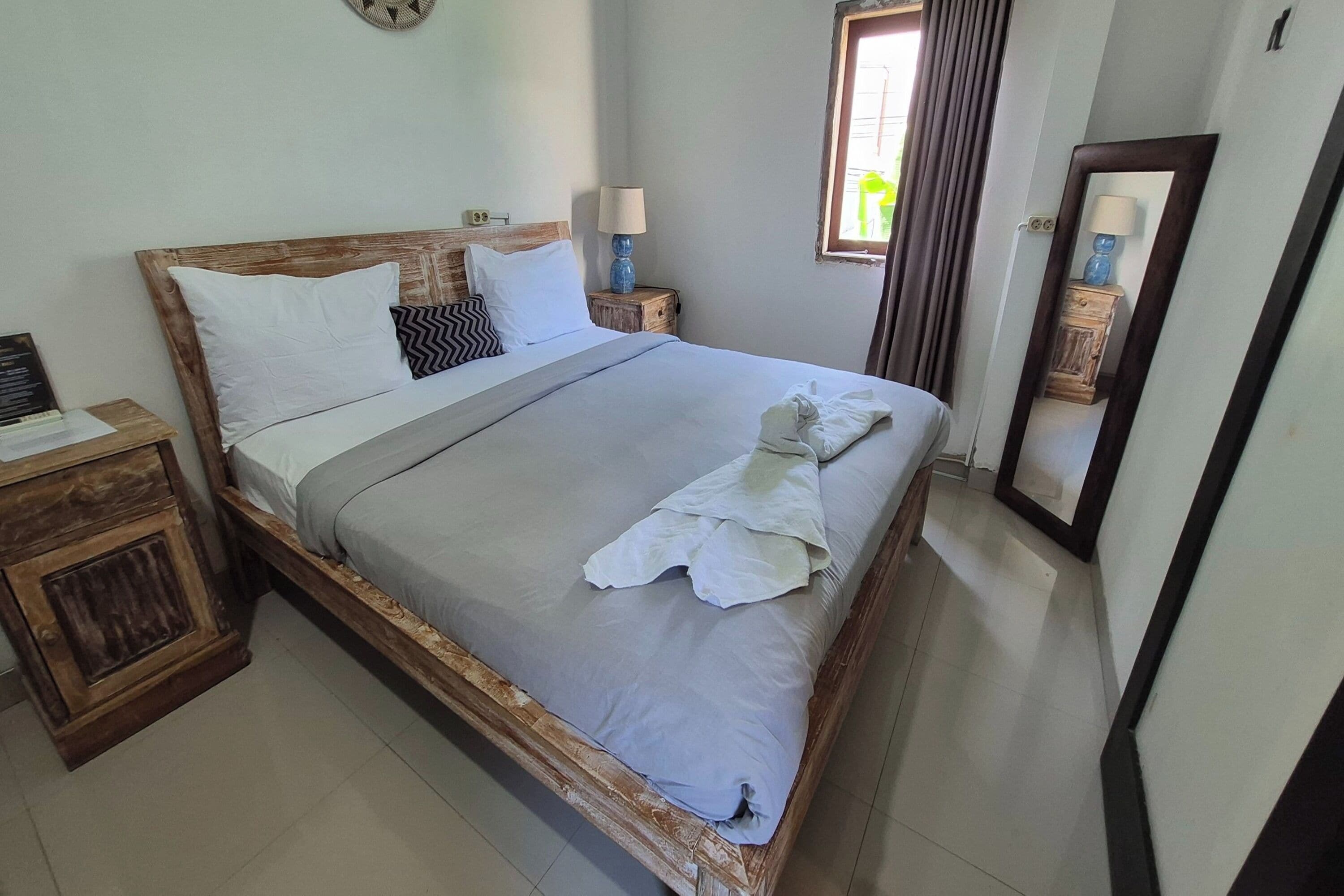 A neatly made bed with white linens and a grey blanket, a wooden headboard, a bedside table with a lamp, and a mirror on the wall.