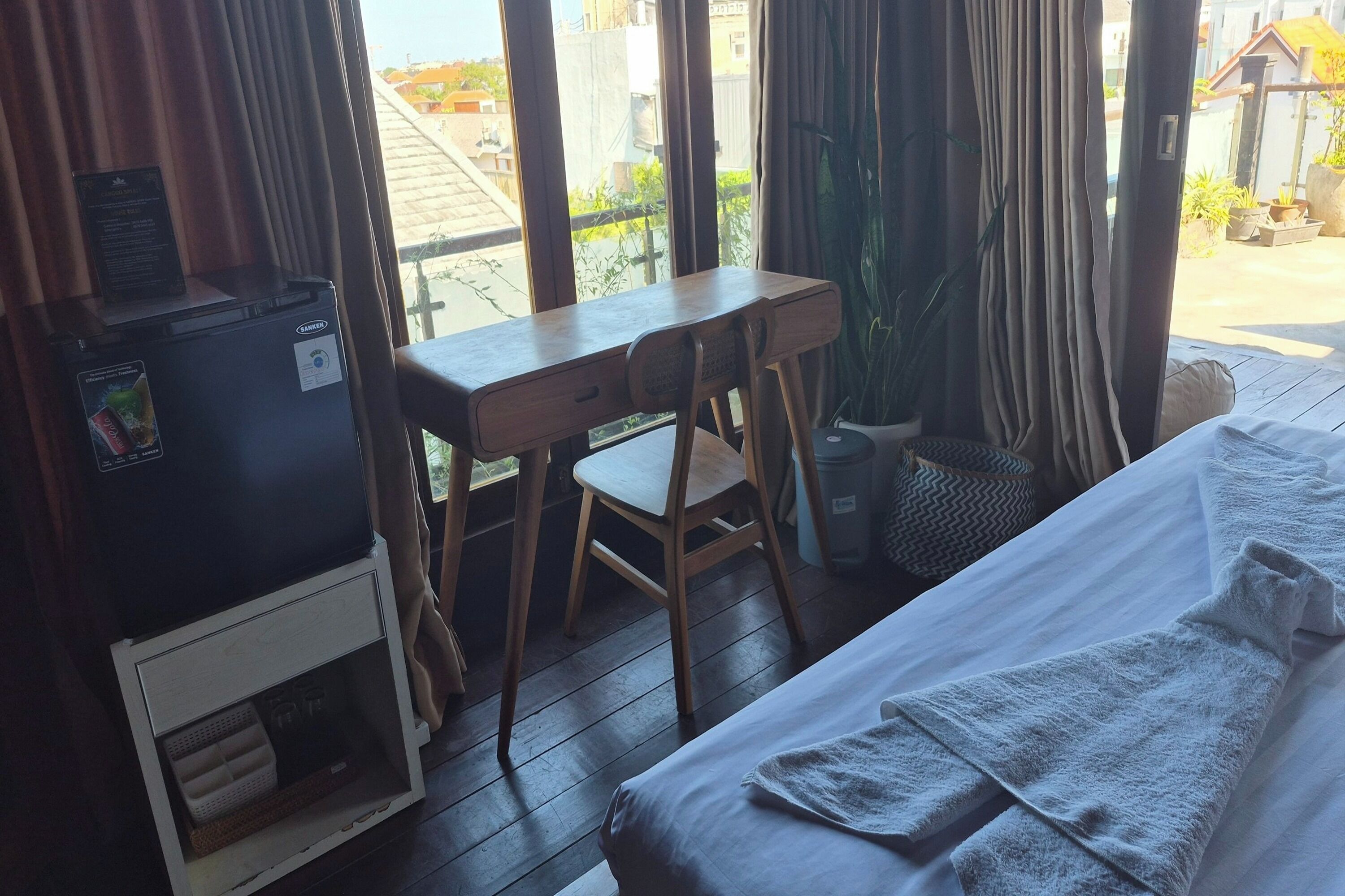 Una habitación con cama, escritorio, silla, un refrigerador pequeño y una ventana con cortinas.
