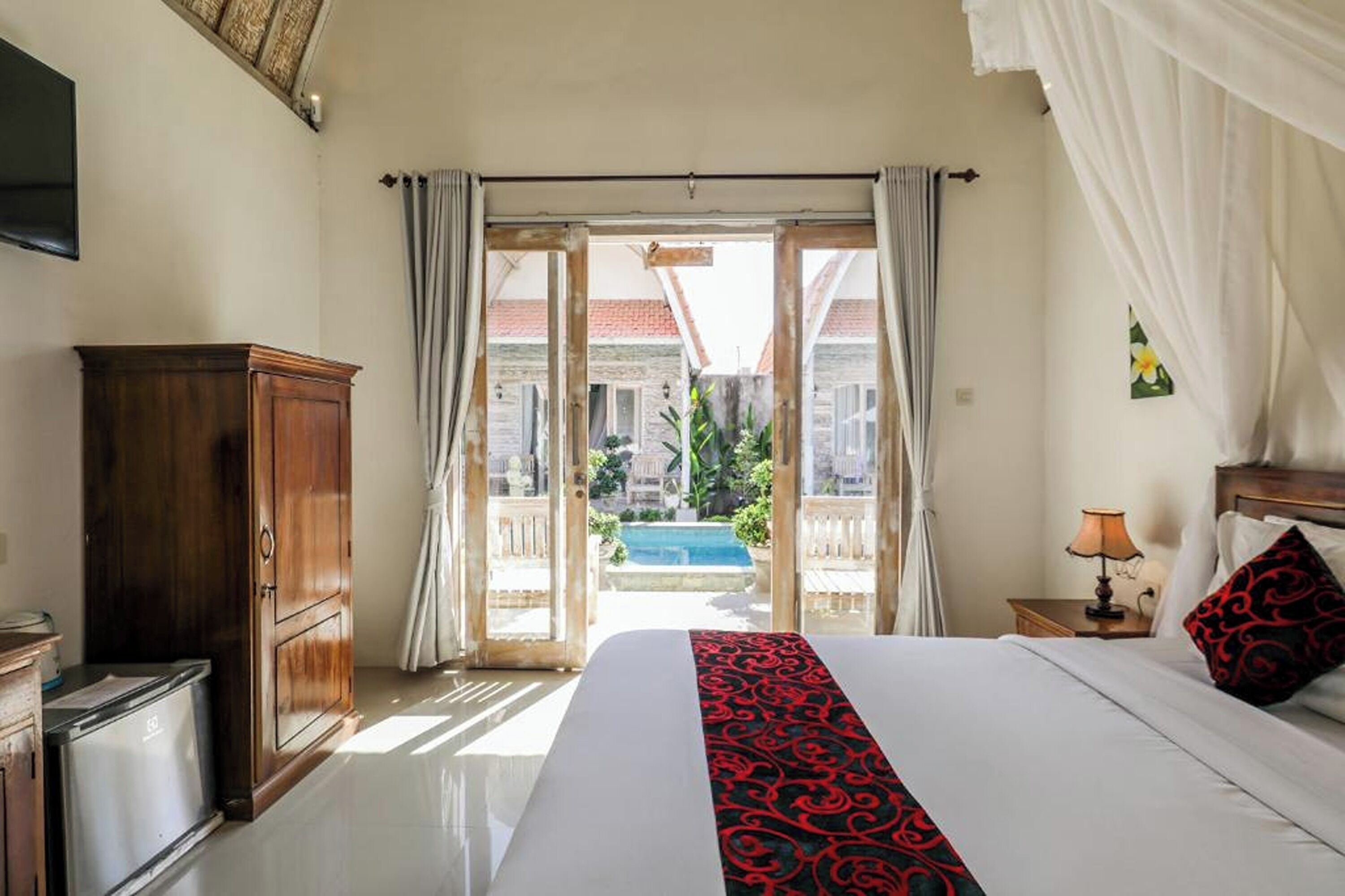 A hotel room with a bed, a wooden cabinet, a desk, a lamp, and a view of an outdoor swimming pool.