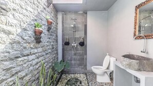 A bathroom with a stone wall, a shower, a toilet, and a sink with a stone basin. - The Box Paradise Bungalow (Penida Island)