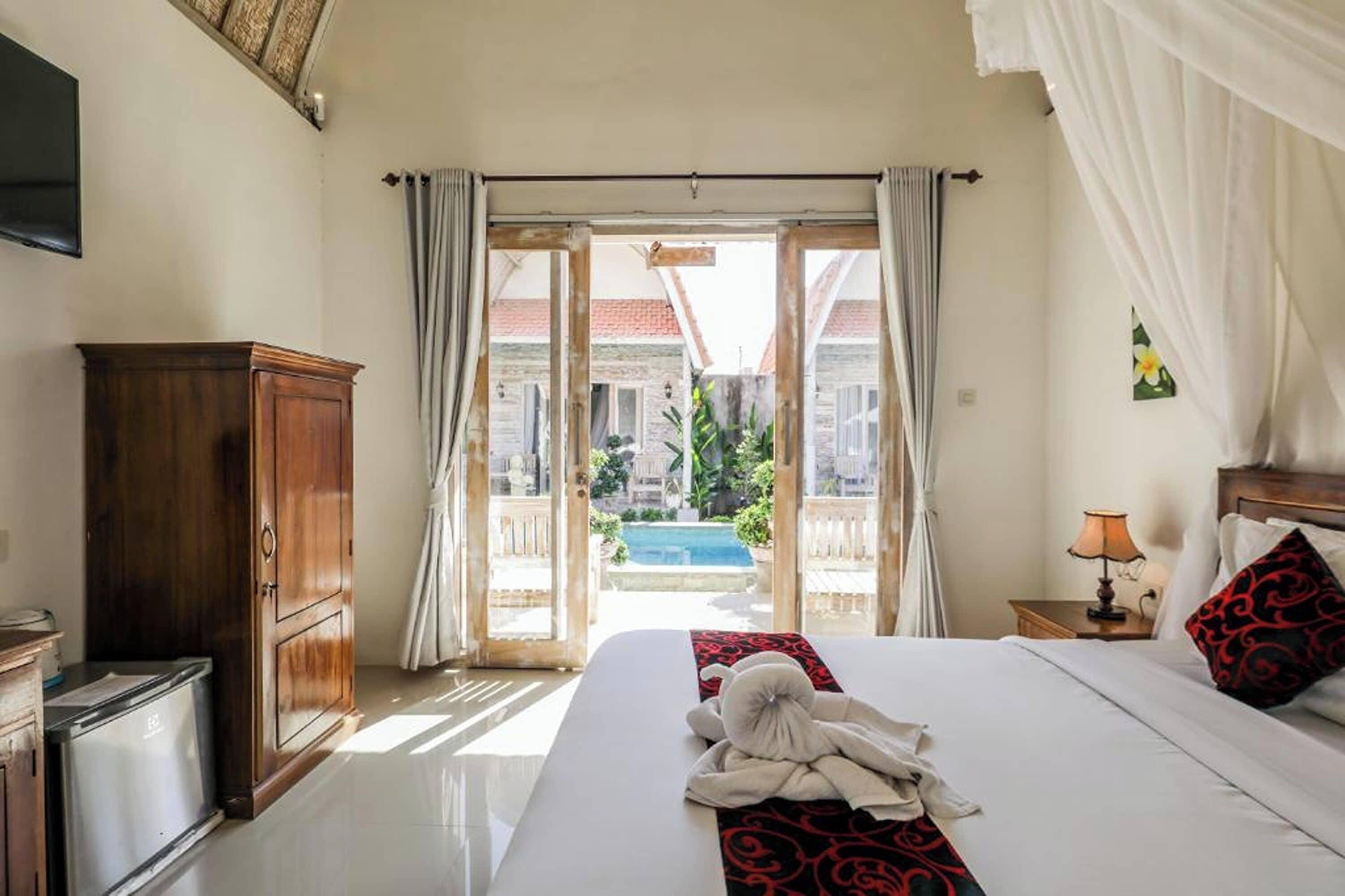 A hotel room with a bed, a wooden cabinet, a nightstand with a lamp, and a view of a swimming pool and buildings outside.