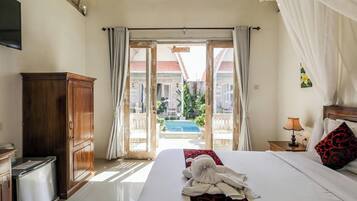 A hotel room with a bed, a wooden cabinet, a nightstand with a lamp, and a view of a swimming pool and buildings outside.