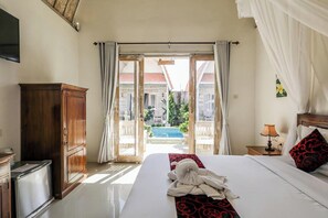 A hotel room with a bed, a wooden cabinet, a nightstand with a lamp, and a view of a swimming pool and buildings outside. - The Box Paradise Bungalow (Penida Island)