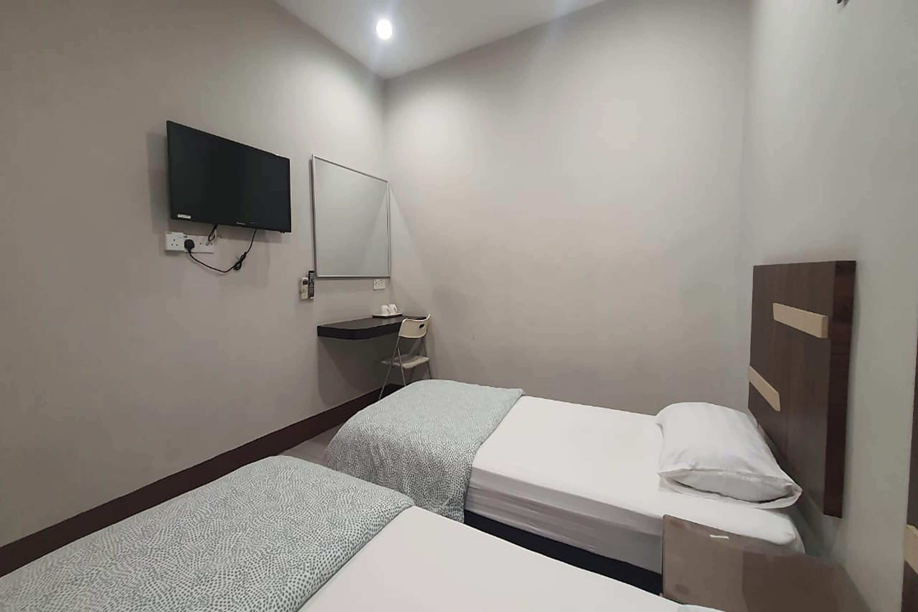 Habitación de hotel con dos camas individuales, un televisor de pantalla plana montado en la pared, un escritorio pequeño con una silla y un espejo sobre el escritorio.