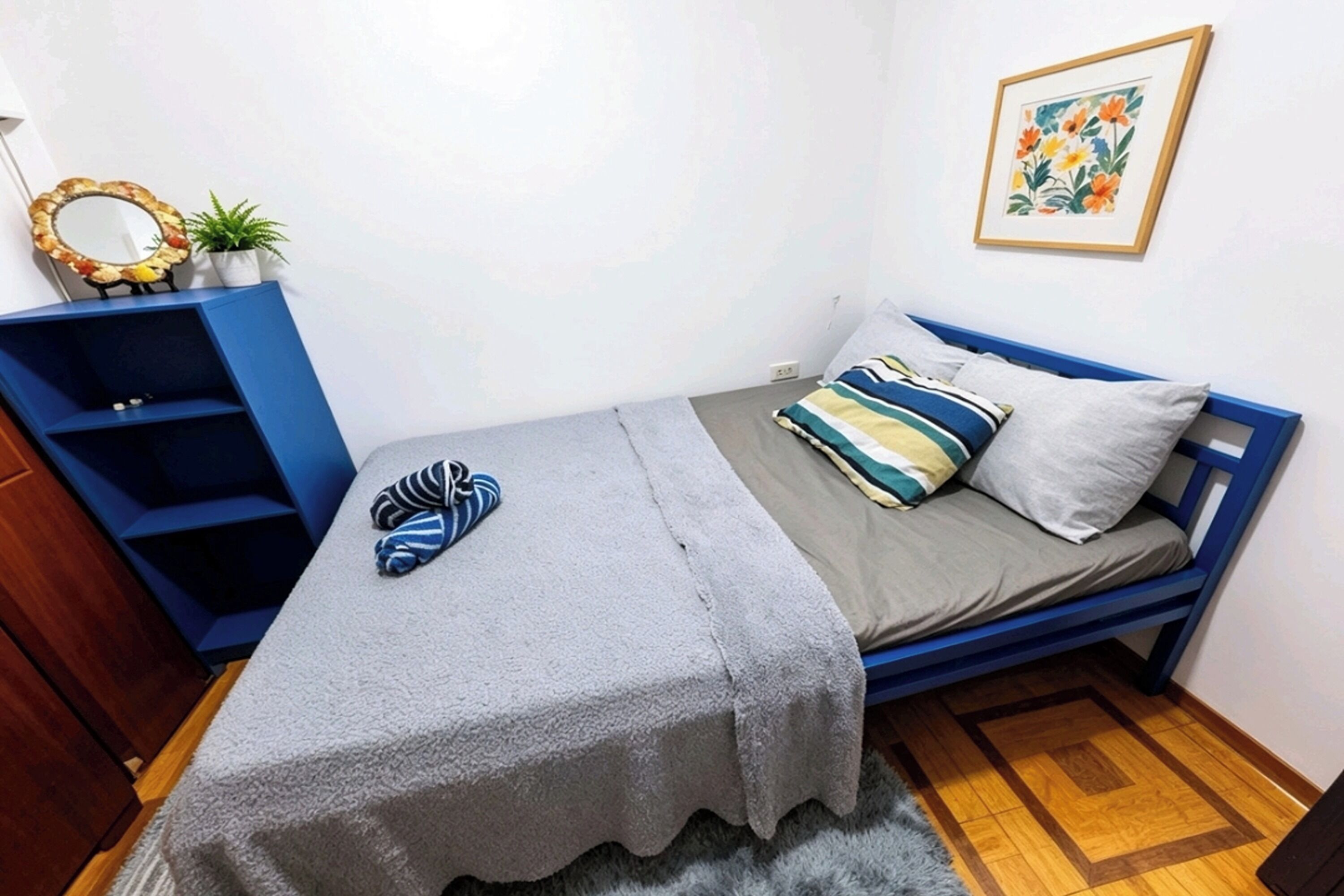 A small, neatly arranged bedroom with a single bed, a blue wardrobe, a small table, and a framed picture on the wall.