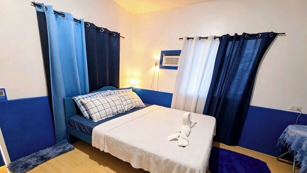 A single bed with a white bedspread and a towel, a blue wall, a small table with a chair, and a blue rug.