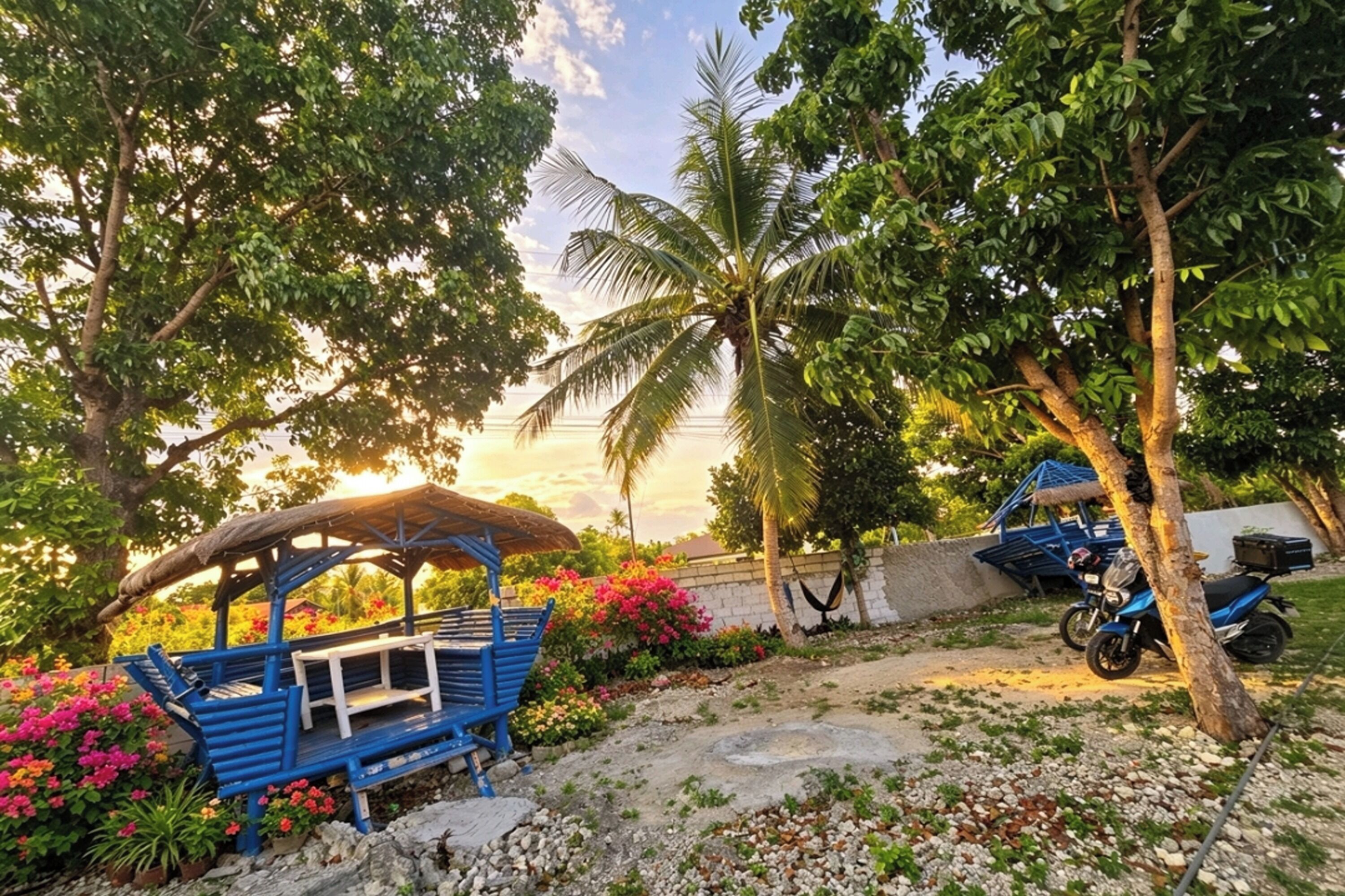A blue outdoor seating area with a thatched roof, surrounded by lush greenery and flowering plants.