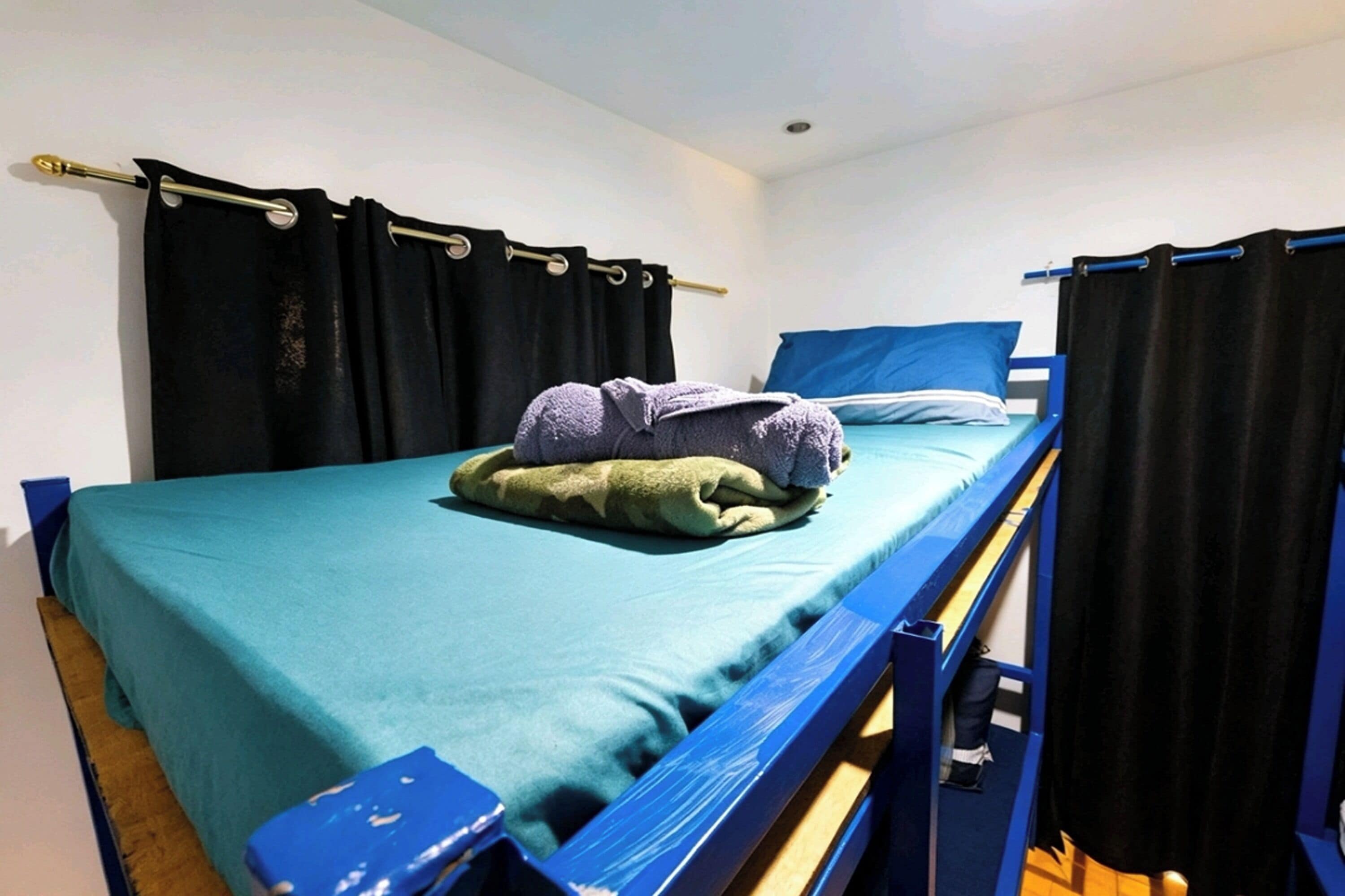 Una cama individual con colchón azul, manta verde y toalla morada.