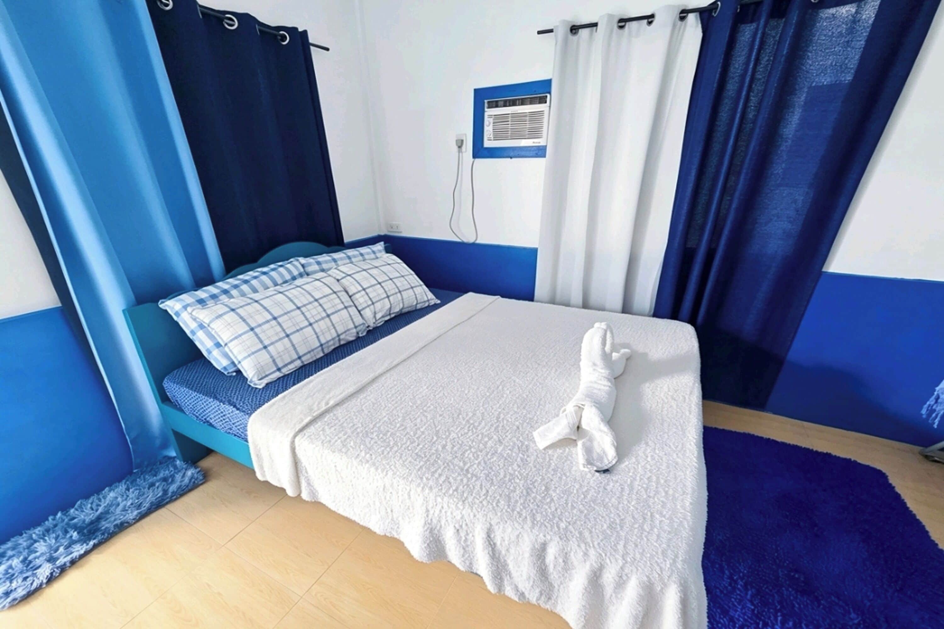 Une chambre à coucher avec un lit, un oreiller à carreaux bleus et blancs, une couverture blanche et un tapis rayé bleu et blanc.