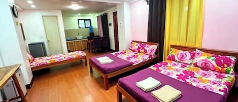 A room with two beds, a desk, a chair, a small kitchenette, and a bathroom.