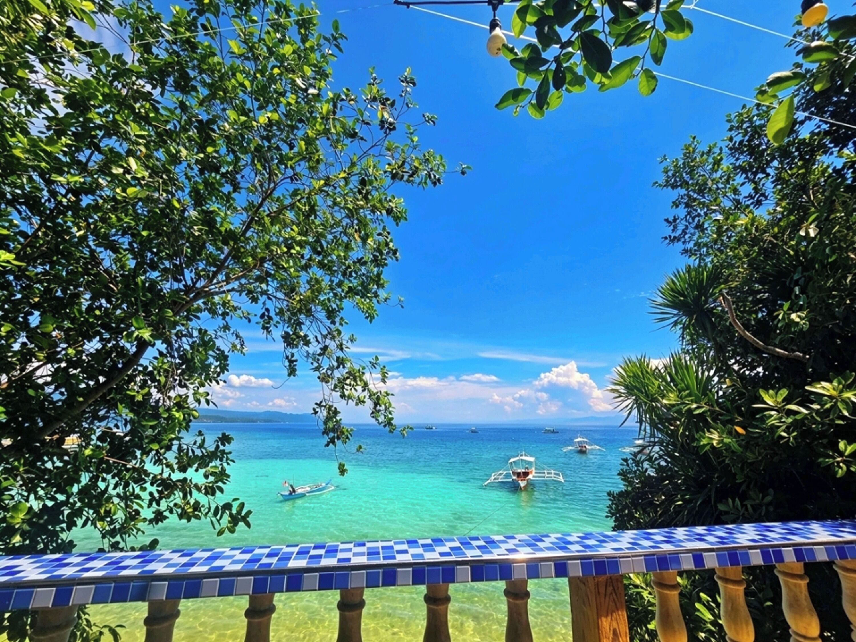 A balcony with a checkered blue and white tile edge overlooking a clear blue sea with boats.