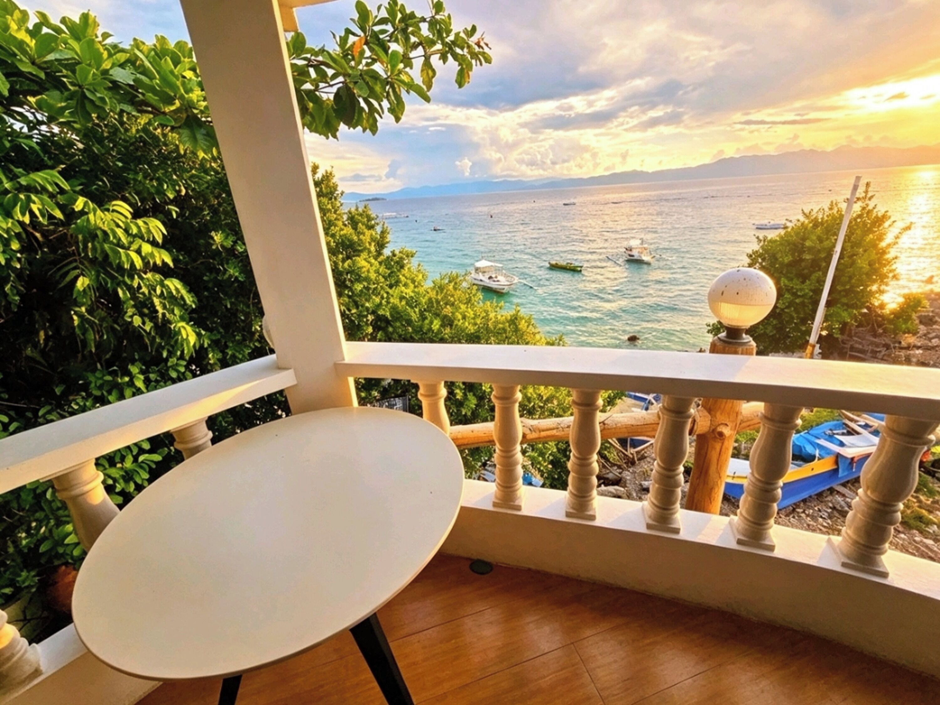 A balcony with a view of the sea and boats, a round table, and a lamp post.