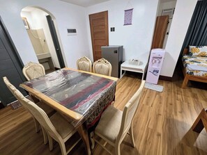 Una habitación con una mesa de comedor, cuatro sillas, una mesita pequeña blanca, un refrigerador y una cama con colcha estampada.