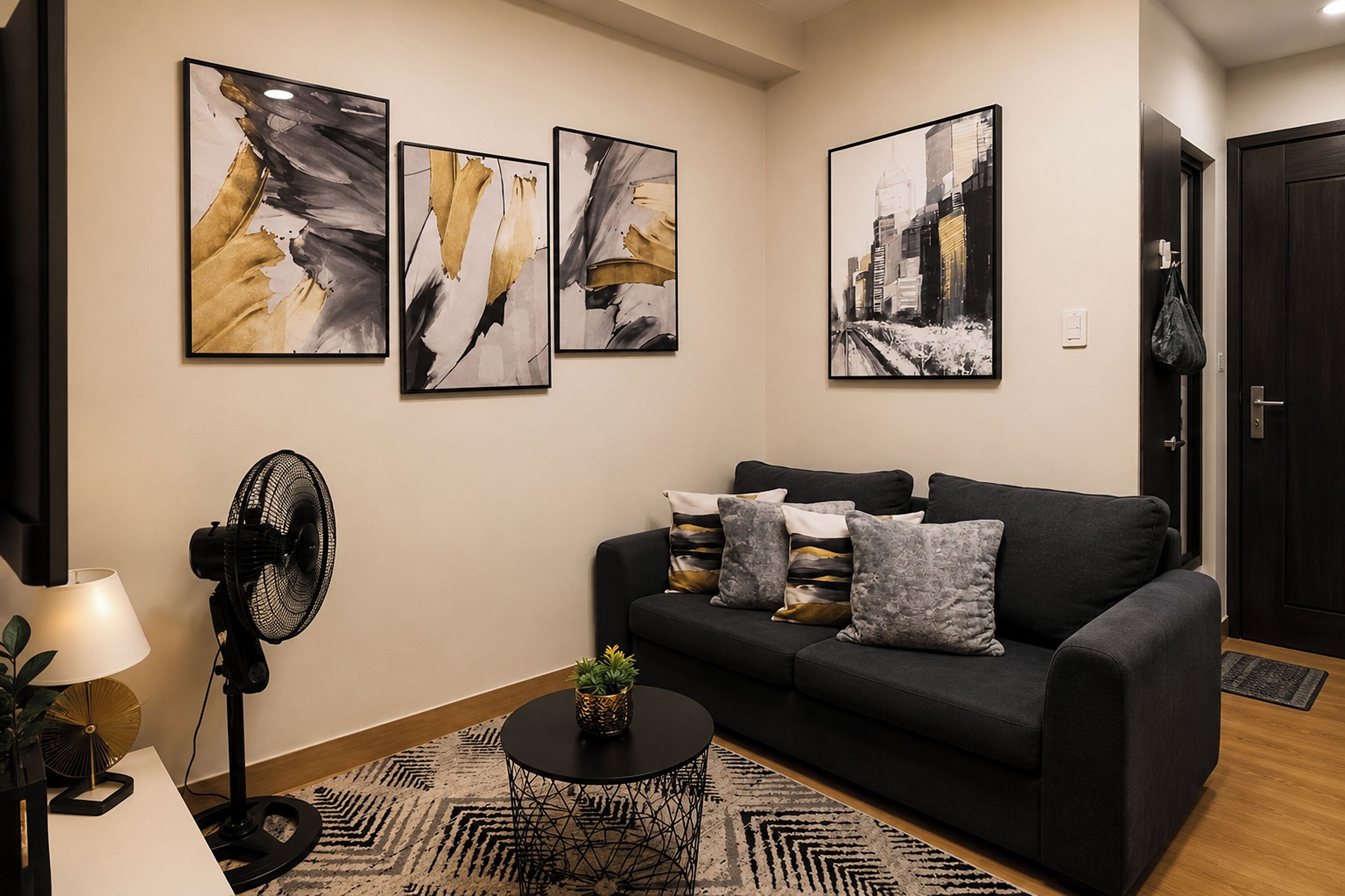 A modern living room with a black sofa, abstract wall art, a standing fan, and a small table with a plant.