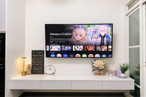 Televisão de tela plana montada em parede branca exibindo um anime com legenda. Abaixo da TV, um console branco com um relógio, uma placa com frase motivacional e um pequeno vaso de planta.