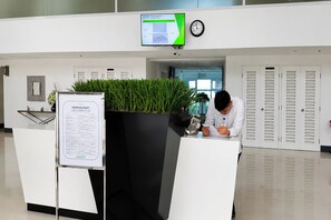 Balcão de recepção com monitor de computador exibindo software de gestão de propriedades, um relógio e área de espera com uma planta e uma placa de identificação.