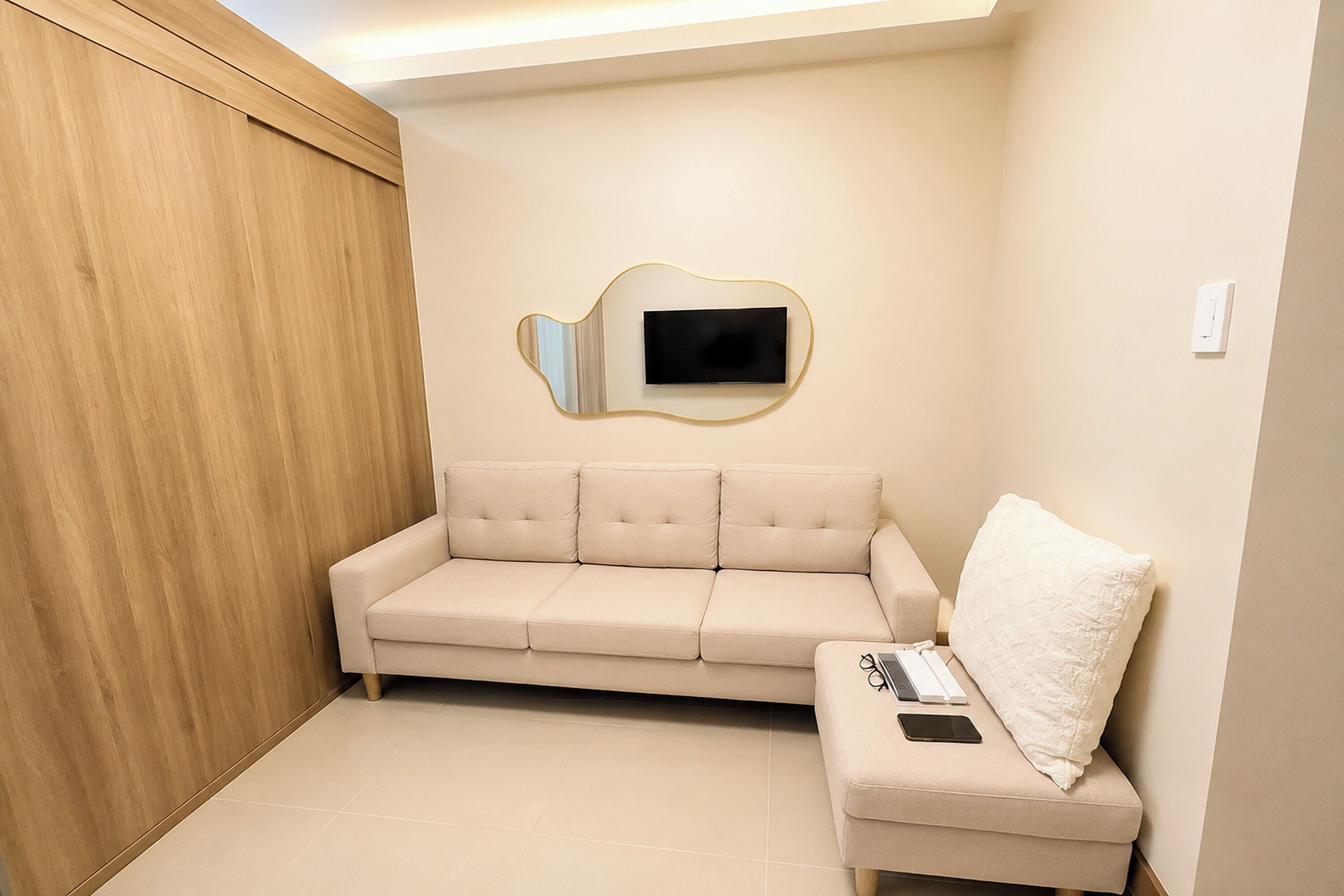 A room with a beige sofa, a small table with a remote control, and a TV mounted on the wall.