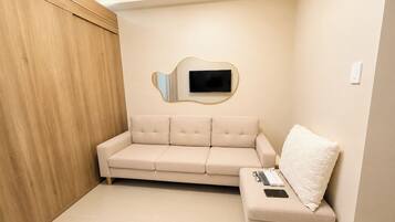 A room with a beige sofa, a small table with a remote control, and a TV mounted on the wall.