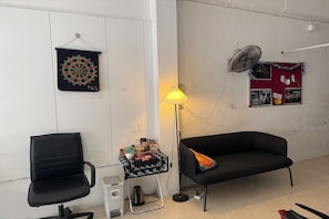 A room with a black office chair, a black sofa, a standing lamp, a small table with a plant, a kettle, and a trash bin. - Sleep Break Hostel Sathon (Bangkok)