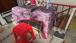 A small dining area with a table covered by a patterned tablecloth, a red plastic stool, and a weighing scale on the floor. - Sleep Break Hostel Sathon (Bangkok)