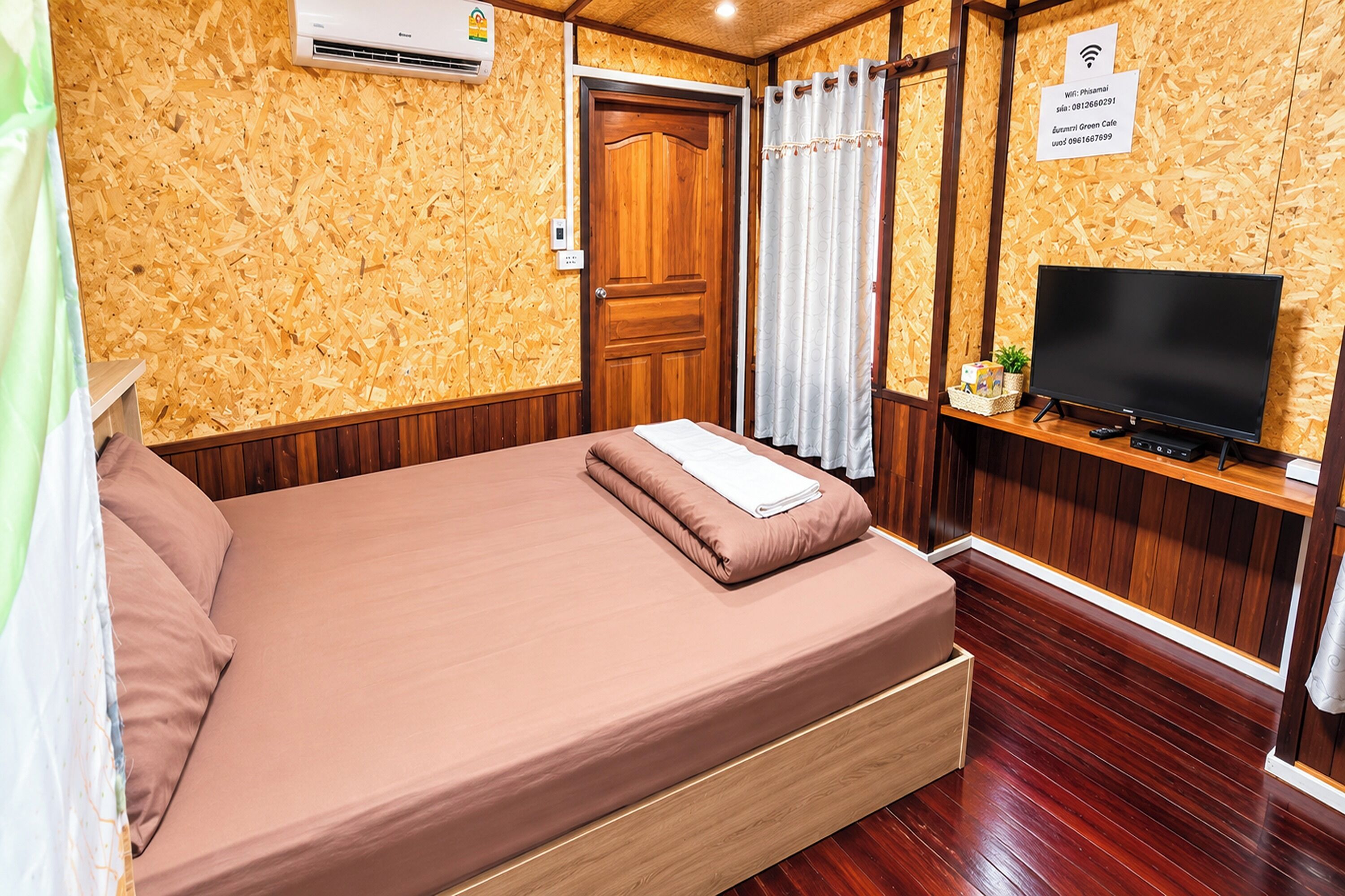 Una habitación con piso de madera, una cama con colchón marrón, un televisor sobre un mueble de madera y una puerta con marco de madera.