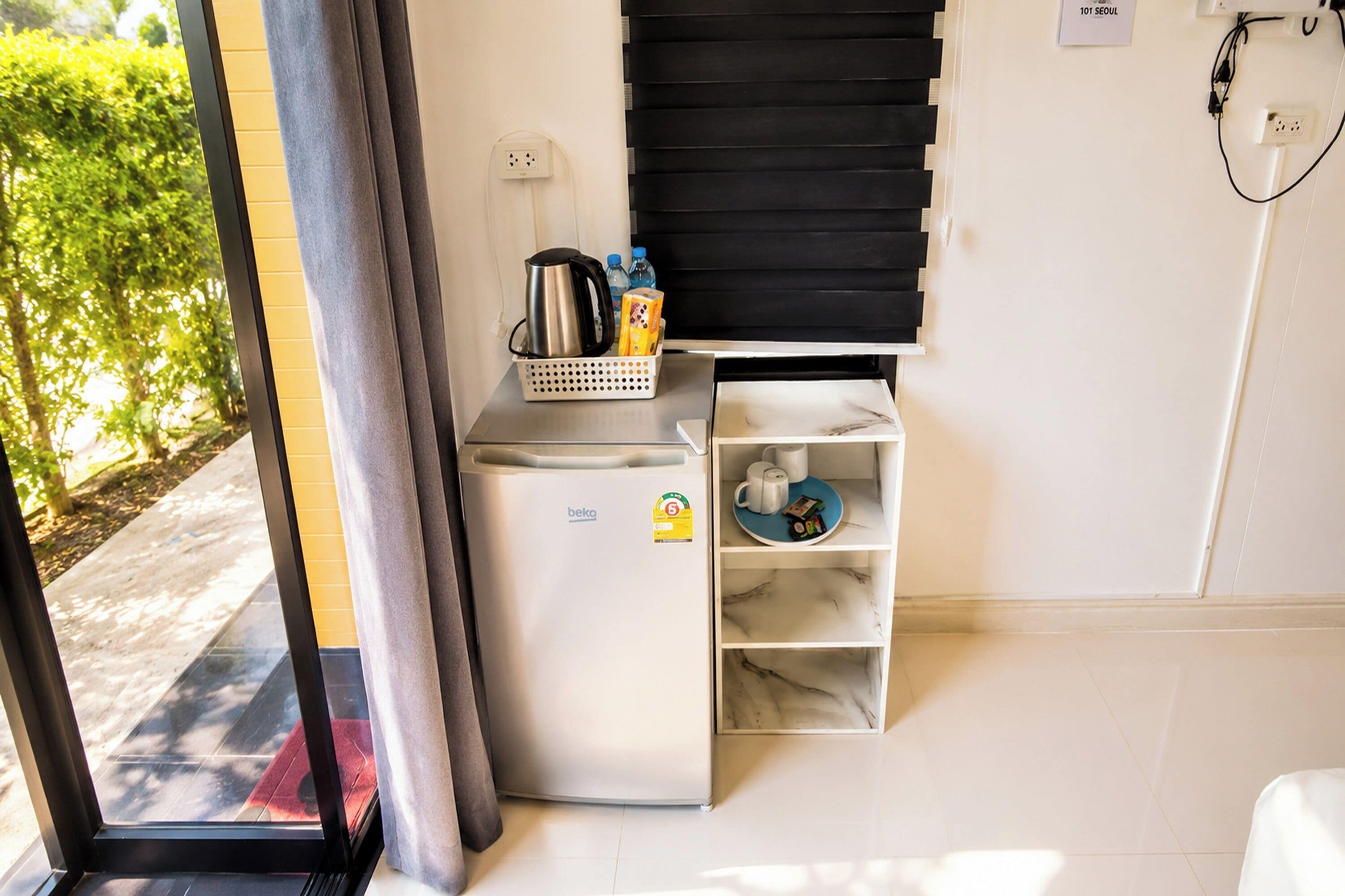 A compact kitchenette with a refrigerator, a coffee maker, and a small shelf with a mug and a bottle.