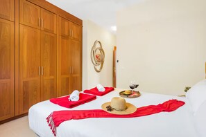 Ett hotellrum med en stor garderob, en säng med röda handdukar och en halmhatt, och ett brickbord med ett glas rött vin.