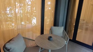 A modern outdoor seating area with a round table and two chairs, large windows with curtains, and a view of trees. - Helena Living Harmony (Varna)