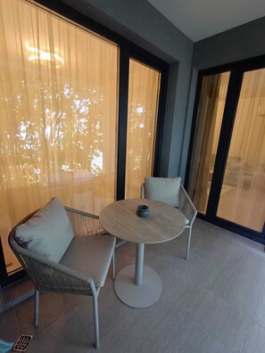 A modern outdoor seating area with a round table and two chairs, large windows with curtains, and a view of trees.