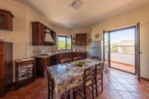 A kitchen with a dining table, wooden cabinets, a refrigerator, and a view through a sliding door. - Hospitality as a Value: ​There are places that keep alive the spirit of those who loved them. (Giardoni)
