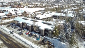 A snow-covered resort with multiple buildings, a parking area with several cars, and a road adjacent to the property. - Wood Fireplace | Hot Tub | Pool in Summer | Arcade Games (Steamboat Springs)