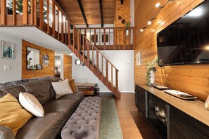 A modern living room with a wooden staircase, a leather sofa, a flat-screen TV, and a dining area. - Wood Fireplace | Hot Tub | Pool in Summer | Arcade Games (Steamboat Springs)