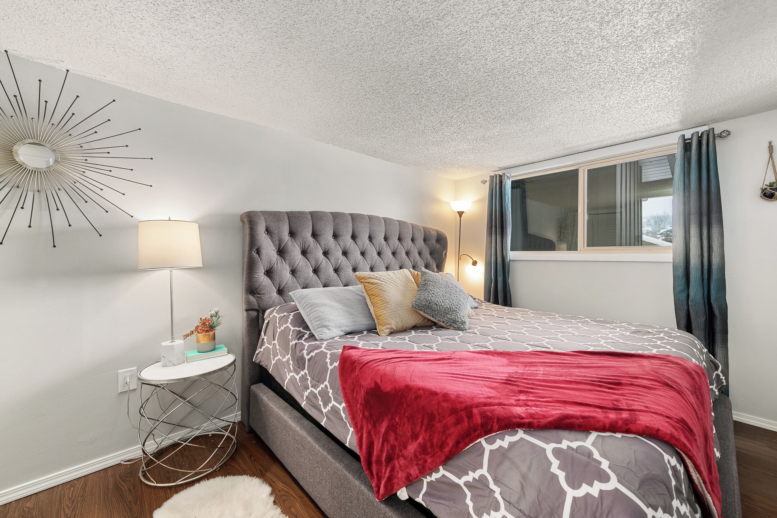 A bedroom with a tufted headboard, a patterned bedspread, a red throw blanket, a side table with a lamp, and a wall-mounted decor piece.