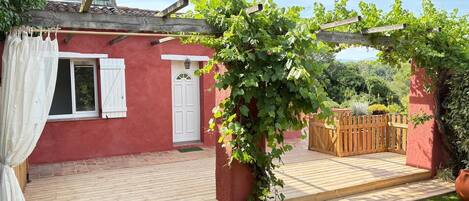 A red building with a wooden deck, a white door, and a pergola with vines.