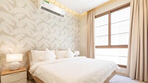 A hotel room with a large bed, a nightstand with a lamp, a wall-mounted air conditioner, and a window with curtains.