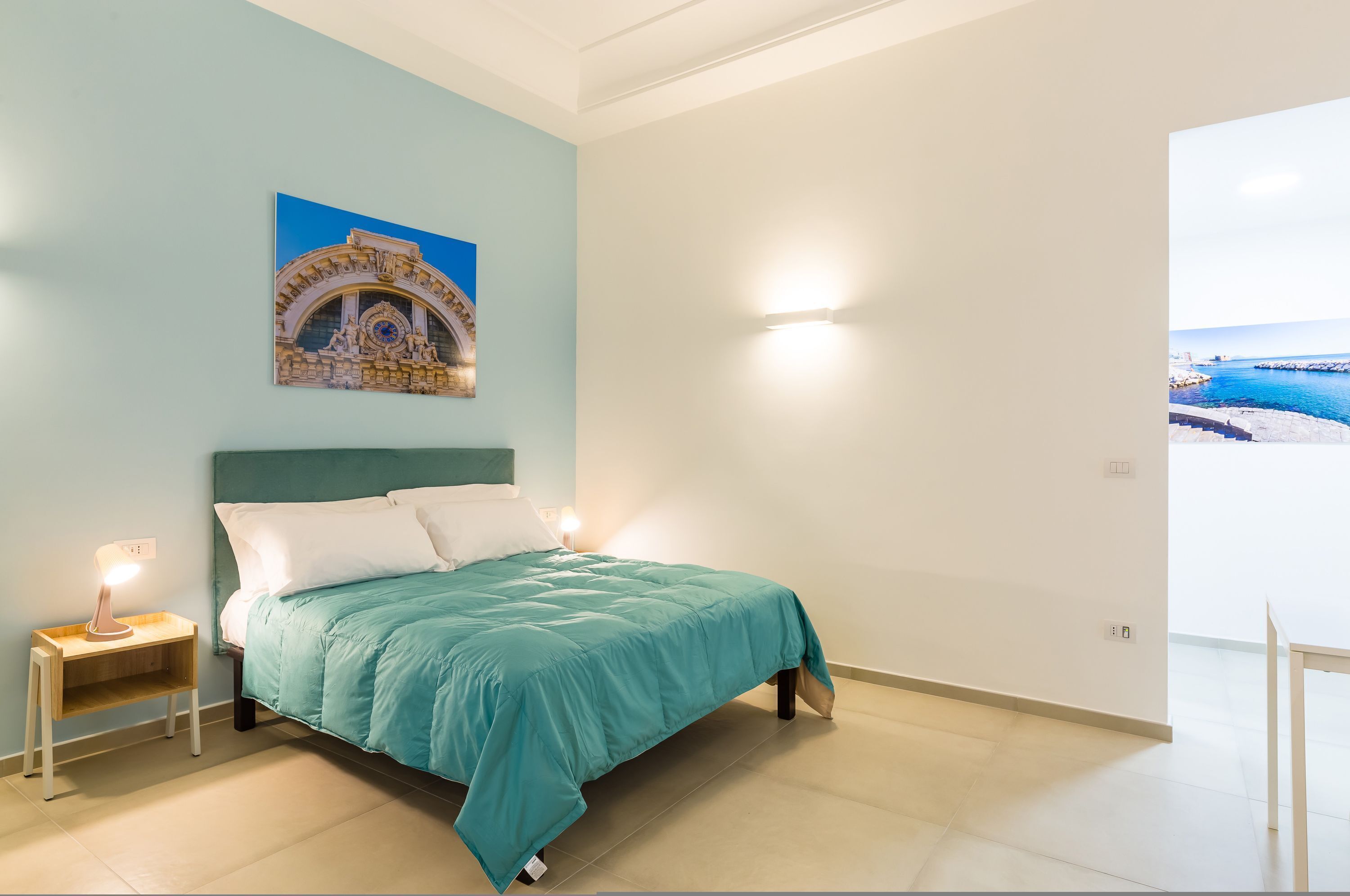 A bedroom with a bed, a nightstand, a wall-mounted lamp, and a painting of a coastal scene.