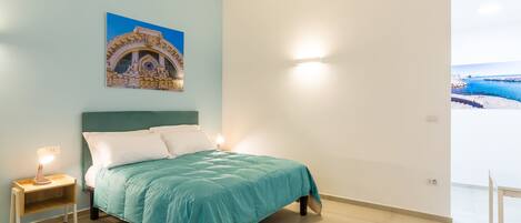 Une chambre à coucher avec un lit, une table de chevet, une lampe fixée au mur, un tableau et une vue sur la mer.