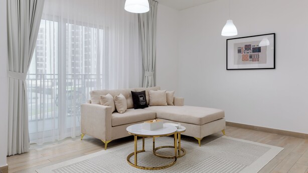 A modern living room with a beige sectional sofa, a round coffee table with gold legs, sheer curtains, and a framed picture on the wall.